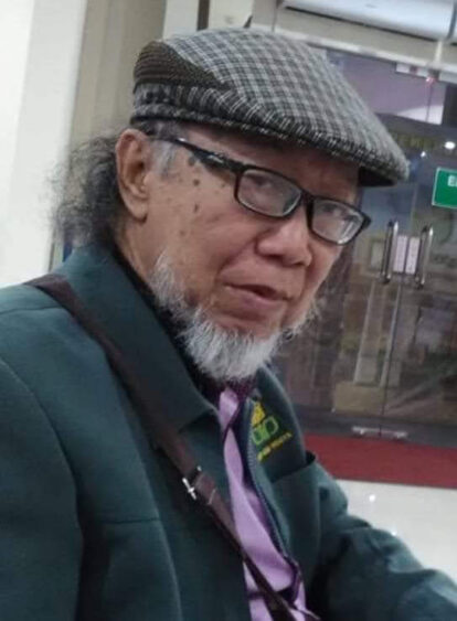 Portrait of an older man with glasses and a gray beard wearing a checkered flat cap, green jacket, and purple shirt indoors.